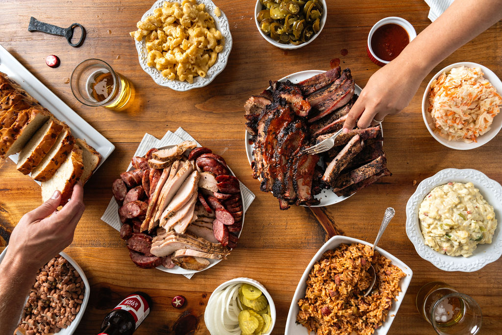 Overhead shot of a variety of meats, sides, and breads, with two hands reaching for different items.