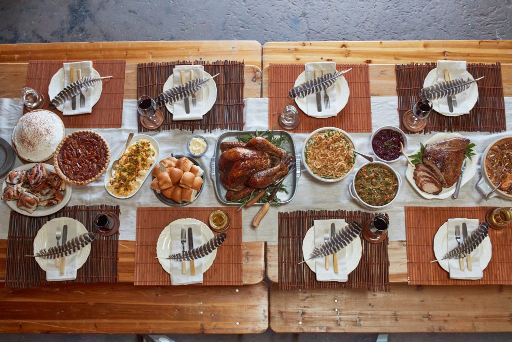Overhead shot of turkey, smoked meats, and all the fixings laid out on a long brown wooden table with white runners, plates, and feathers at each setting.