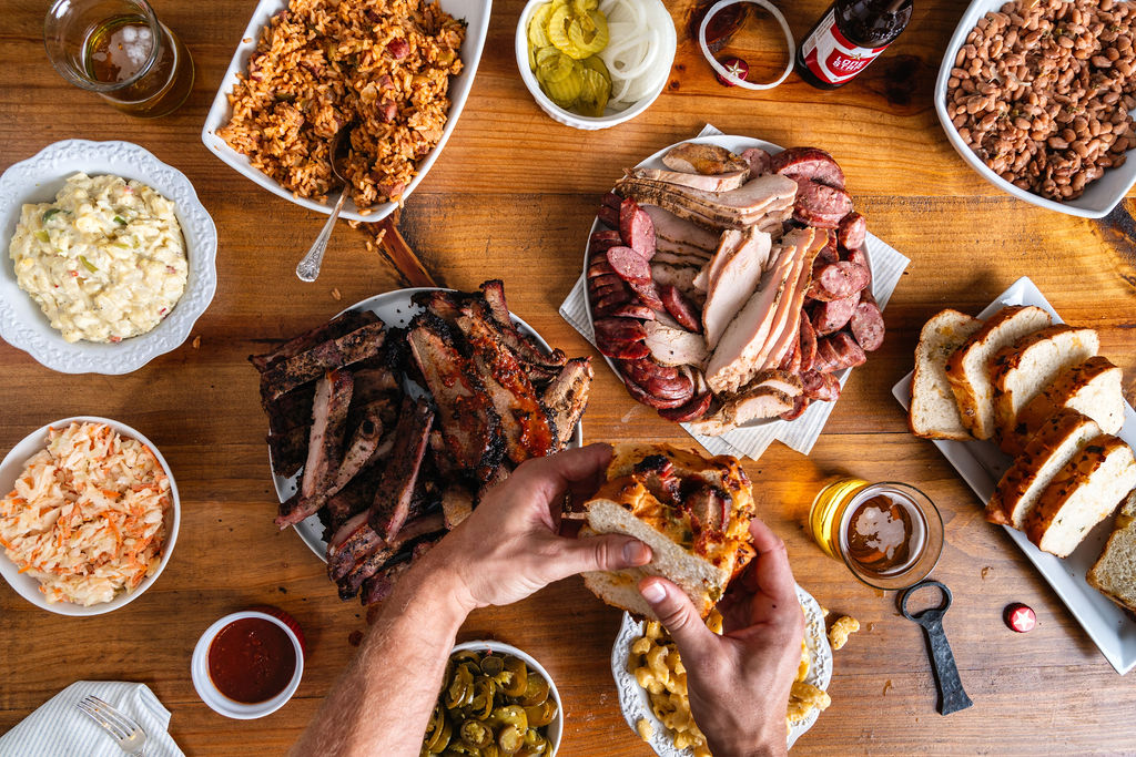 Man holding brisket sandwich over a party pack including ribs, sliced sausage and turkey, potato salad, coleslaw, jalapenos, sliced bread, beans, mashed potatoes, and rice.