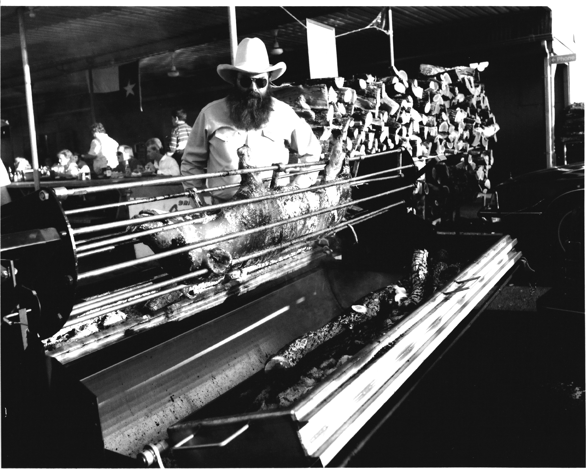 Jim Goode wearing a Stetson and sunglasses, watching a hog being grilled on a BBQ spit.