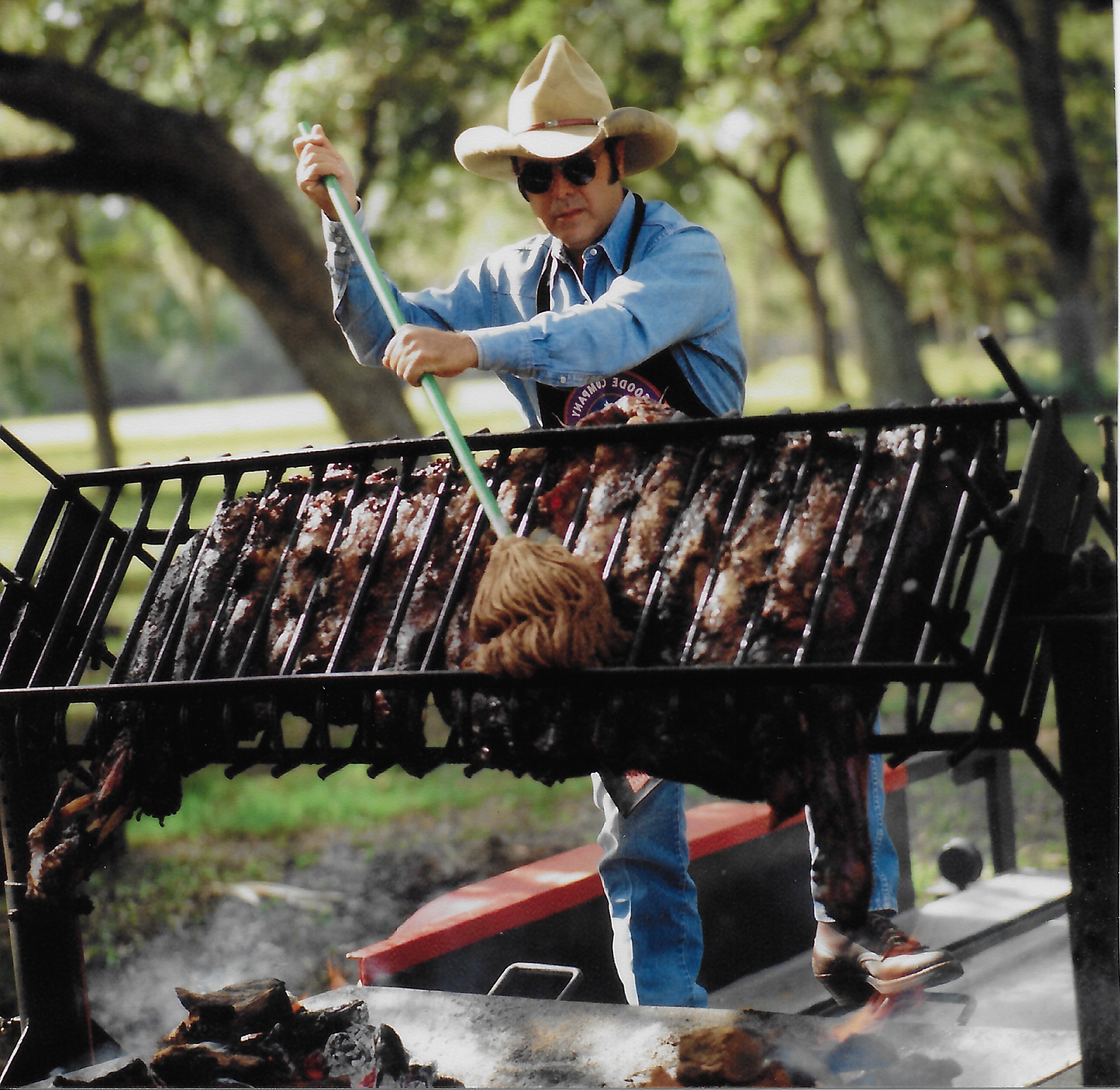 Restaurant founder Jim Goode basting a large rack of ribs outdoors, wearing an apron, sunglasses, and a Stetson.