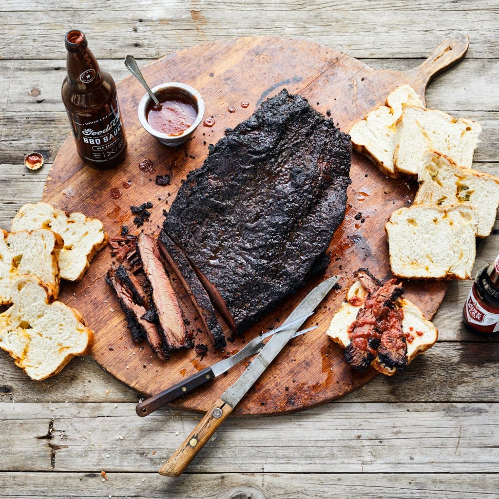 Overhead photo of sliced brisket lying on a wooden cutting board next to a carving knife and fork, sliced jalapeno cheese bread, and a serving of Goode Co. traditional BBQ sauce.