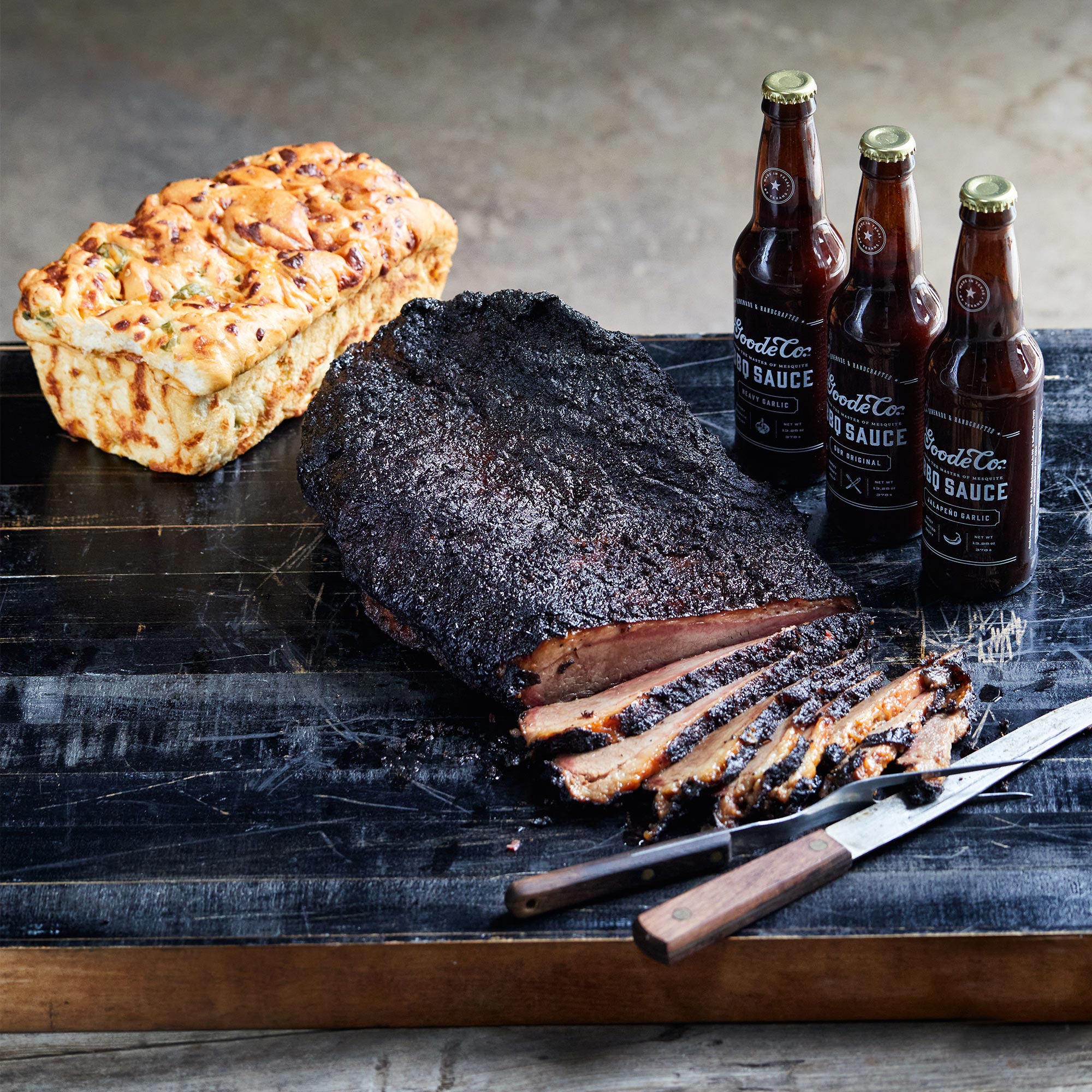 Freshly barbequed brisket on the table with a loaf of jalapeno cheese bread and three bottles of sauce.