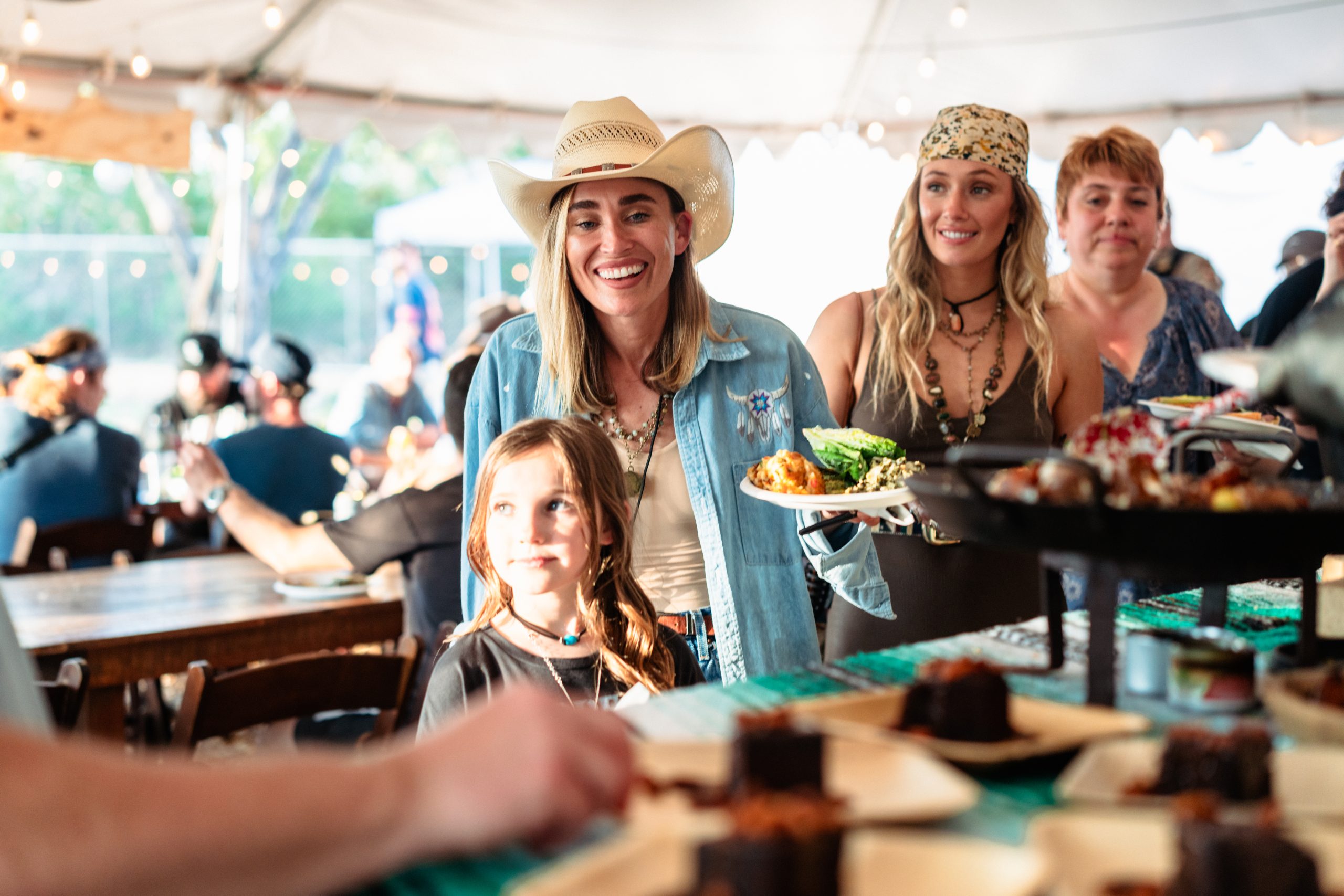 A young blond-haired girl and two blond-haired women wearing stetsons, lined up at a BBQ, each with a hand holding a plate full of food.