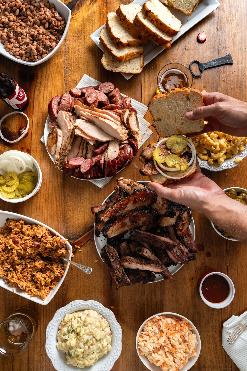 Overhead picture of plates of food with a pair of hands assembling a jalapeno cheese bread, brisket sandwich with onions and pickles.