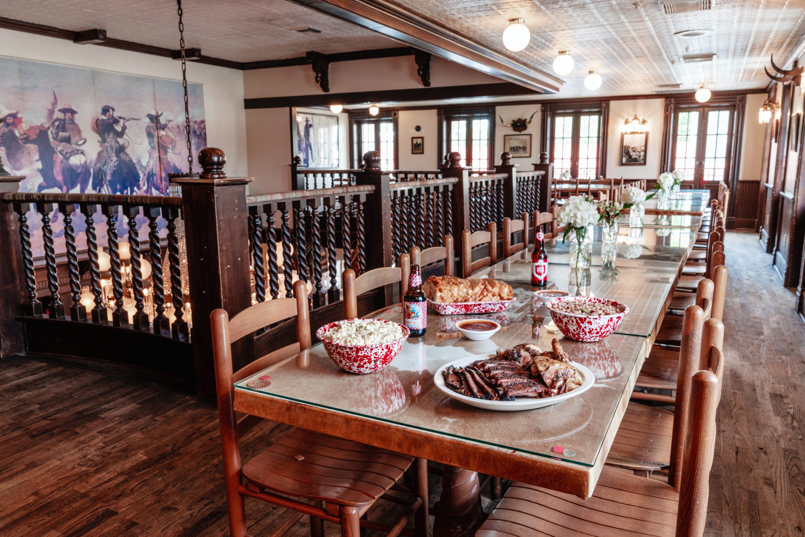 Long dining room table at one of the Goode restaurants featuring a large bowl of potato salad, a selection of BBQ’d meats, beans, beer, and sliced bread.