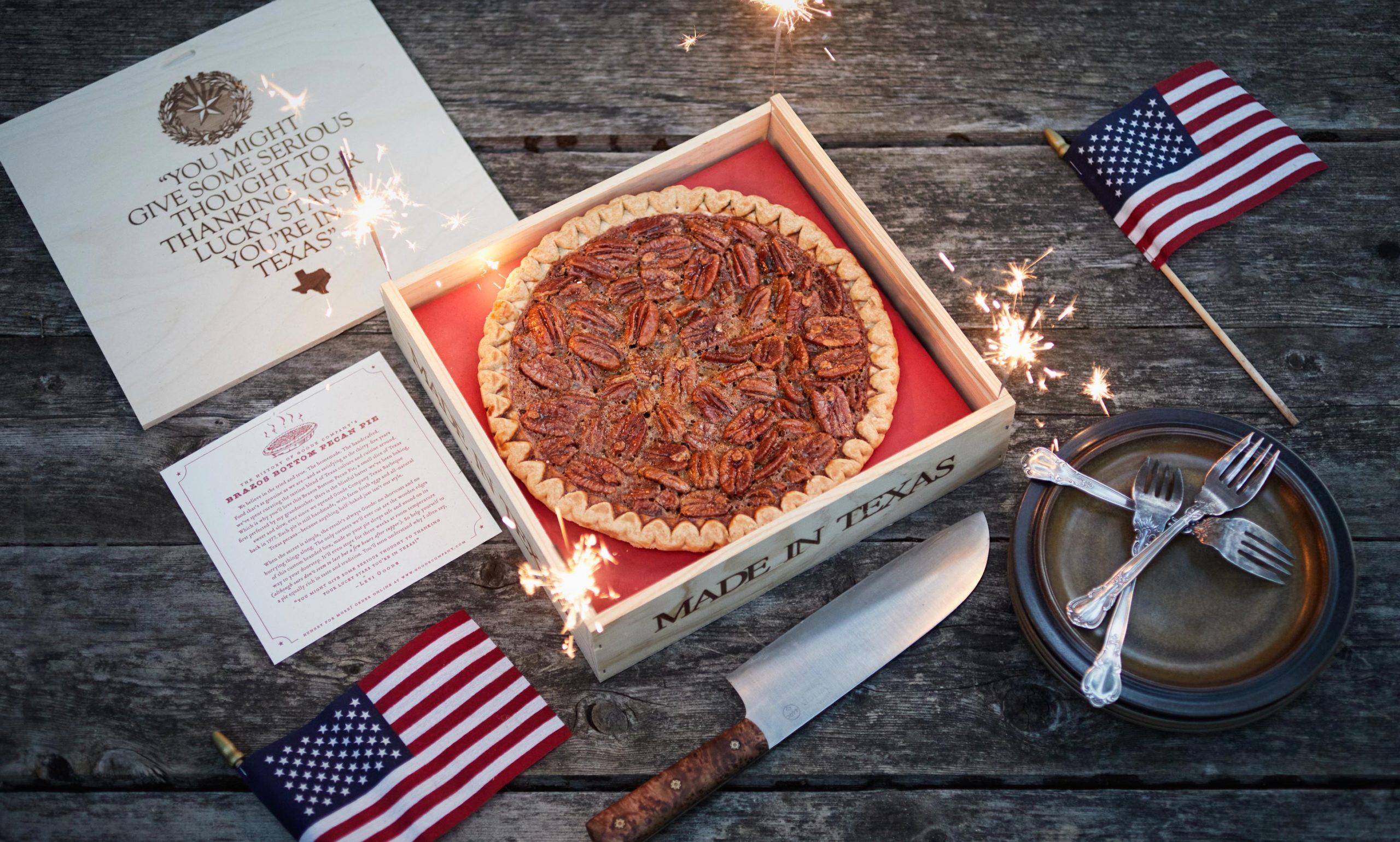 Close-up of pecan pie in a box, surrounded by U.S. flags, sparklers, and forks lying on a plate.